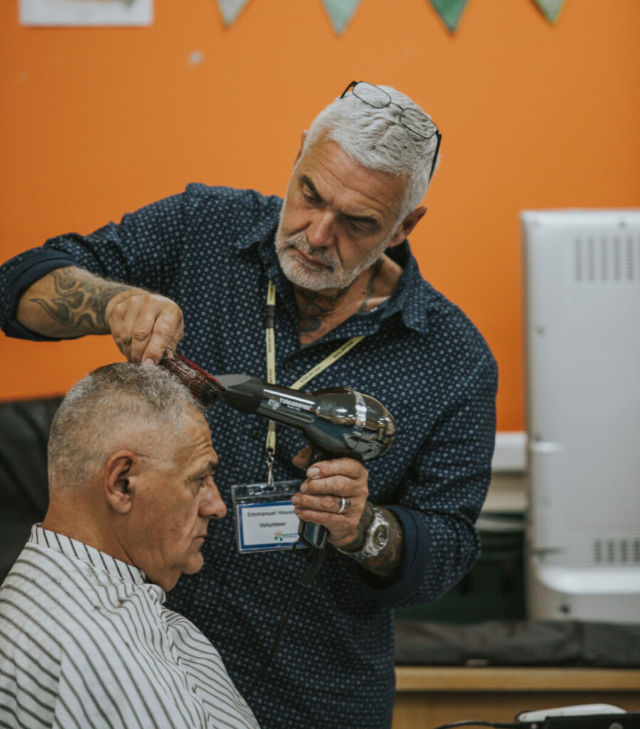 Emmanuel House's Volunteer Barber using a hairdryer to dry and style a service user's hair.