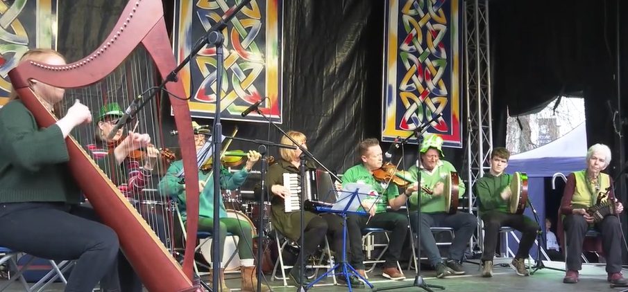 Musicians from the Irish Music Centre sat on stage dressed in green playing a rang of instruments including a harp, accordion, violin, and percussion instruments