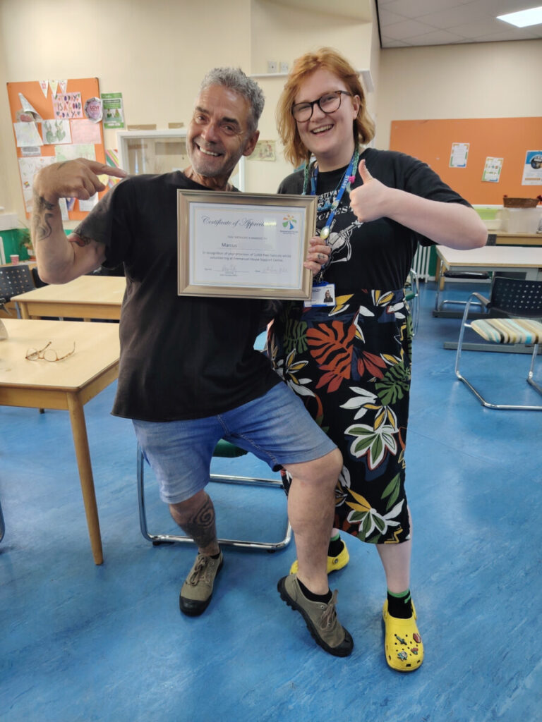 Marcus holding a framed certificate, celebrating his 1,000th haircut during his volunteering at Emmanuel House